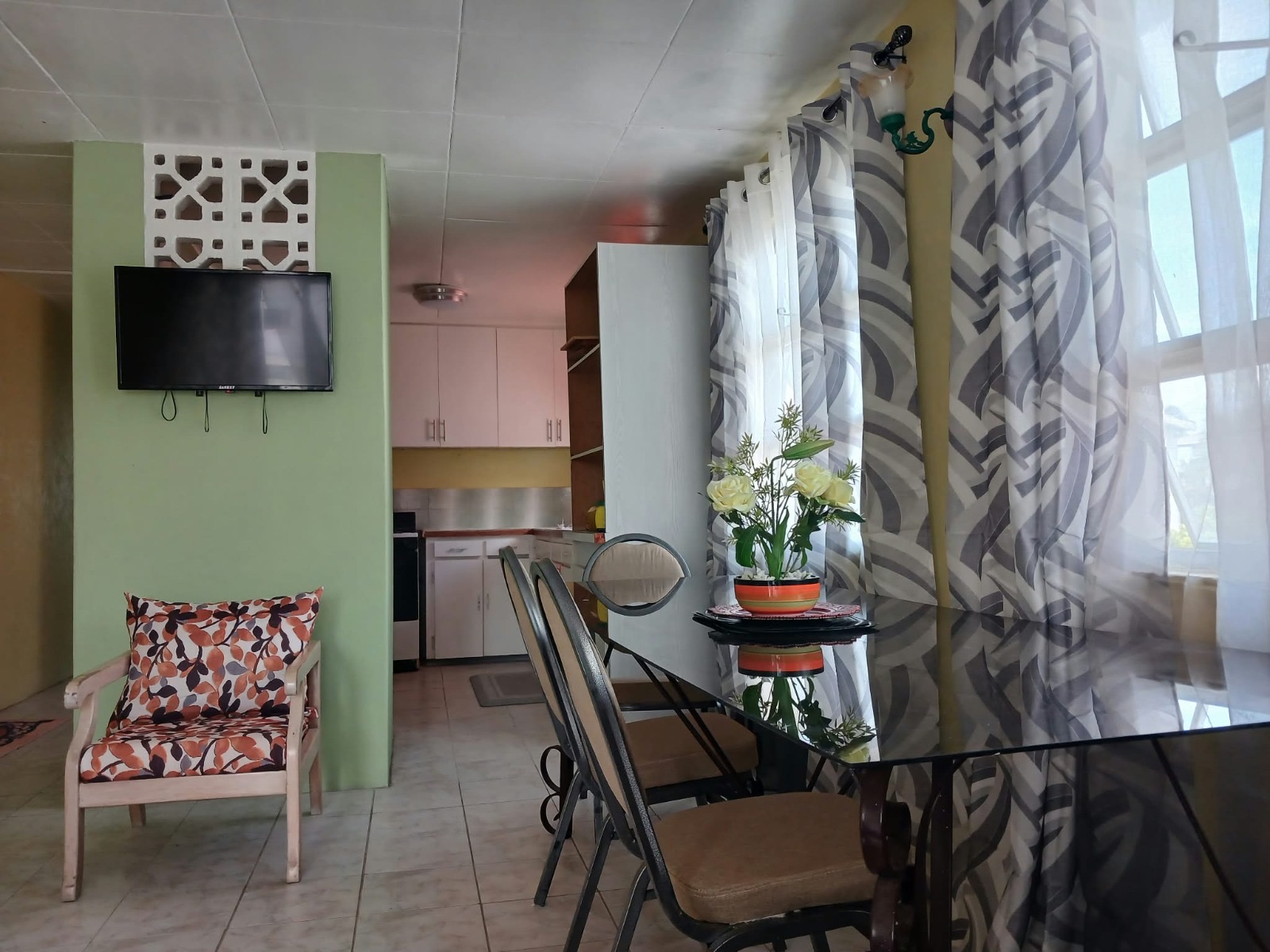 Apartment 3 — living and dining room with wall-mounted TV on a green breeze-block wall, looking into the kitchen